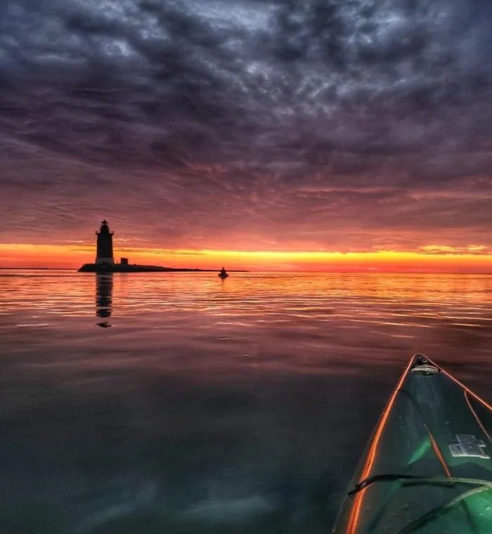 Lighthouse silhouette at sunset over calm waters with a kayaker in the foreground, reflecting the vibrant orange and purple sky.