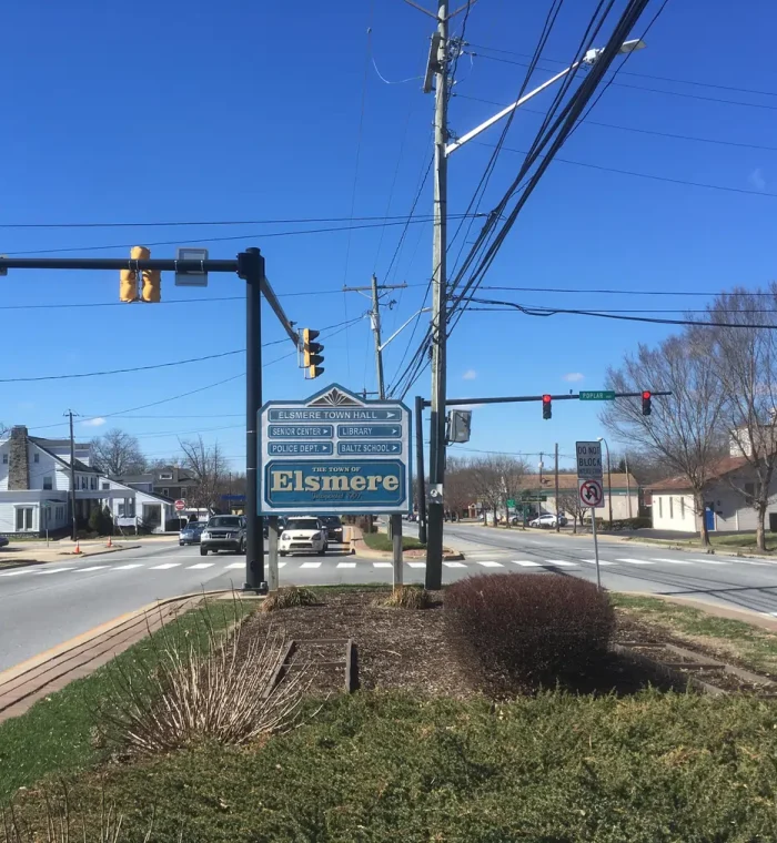Elsmere town sign at Poplar Avenue, displaying local institutions like Town Hall, Library, and Police Department, with clear blue sky and traffic signals.