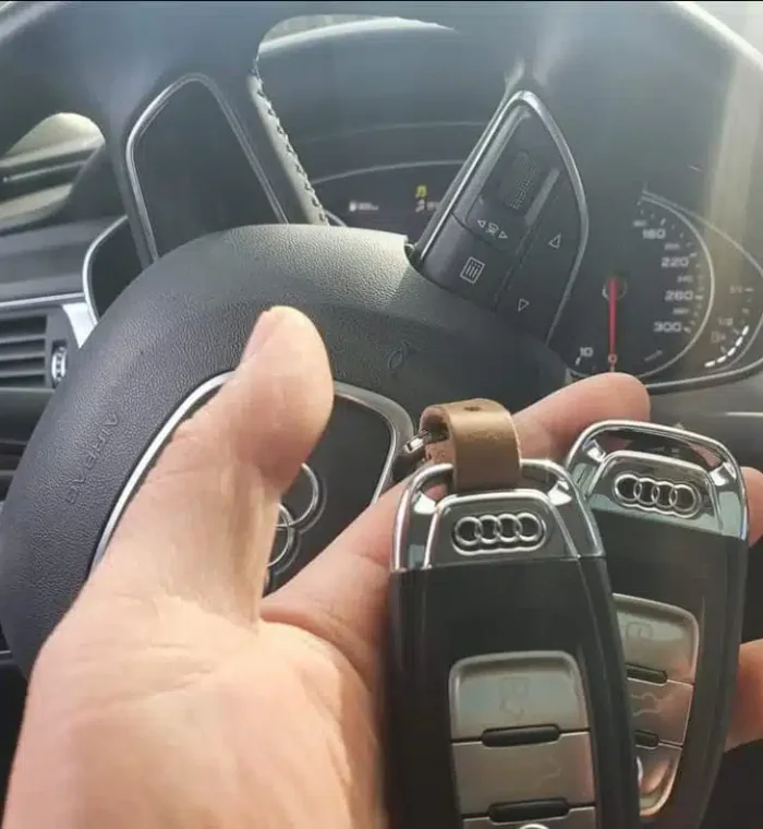 Hand holding two Audi car keys in front of a vehicle's steering wheel and dashboard, illustrating automotive locksmith services and car key replacement by Nova Locksmith.